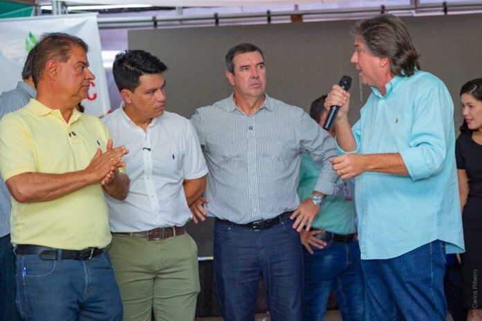 Four men standing on a stage; the man in a light blue shirt is speaking into a microphone while the others listen.