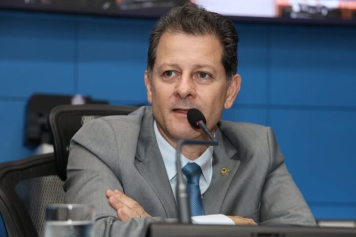 Man in a gray suit speaks into a microphone at a panel, with a blue background behind him.