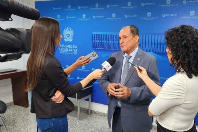 Senator discusses policy with three reporters in a blue legislative backdrop during a press interview.