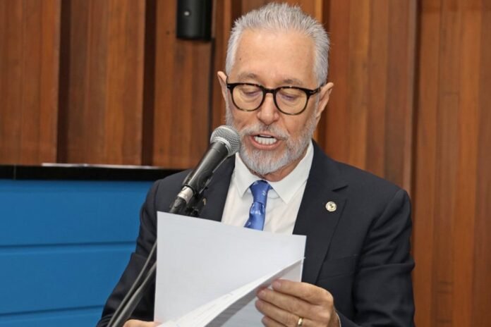 Man in a dark suit and blue tie speaks into a microphone while reading from a stack of papers in a formal setting.
