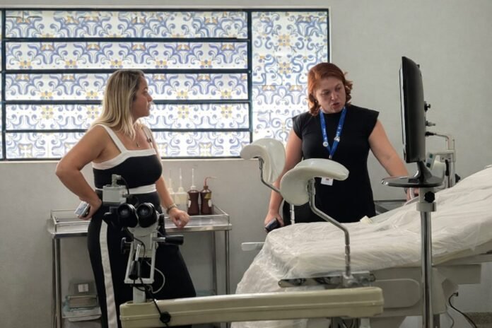Two healthcare workers stand by an exam bed in a hospital room, discussing as medical equipment sits on a nearby cart and a patterned window is in the background.
