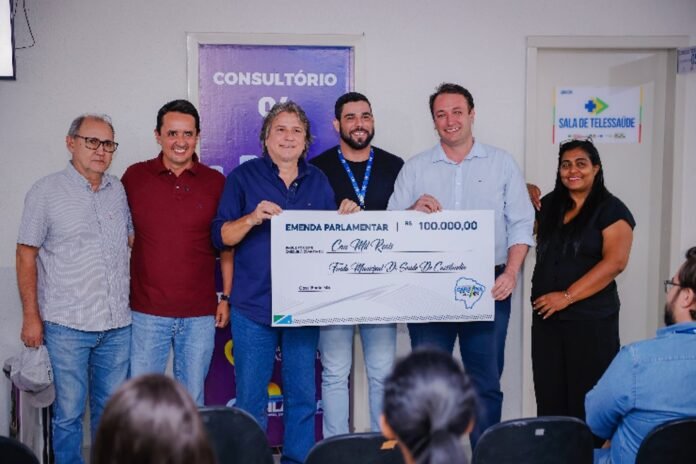 Six people pose in a clinic holding a large ceremonial check for R$100,000 during a parliamentary grant ceremony in healthcare setting.