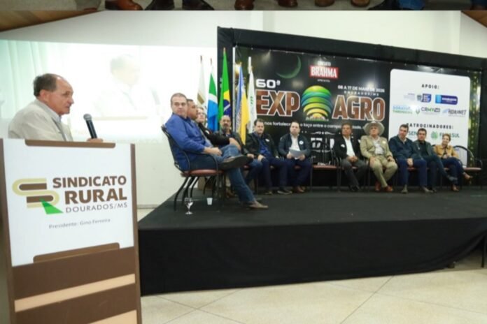 Speaker at a podium addresses a panel of men seated on a stage, with flags and an Expo/Agro banner behind them at a Sindicado Rural event in MS, Brazil.