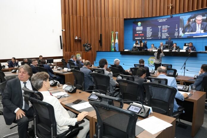 Parliament chamber with officials seated at desks facing a dais and a large blue display board showing agenda and scores.