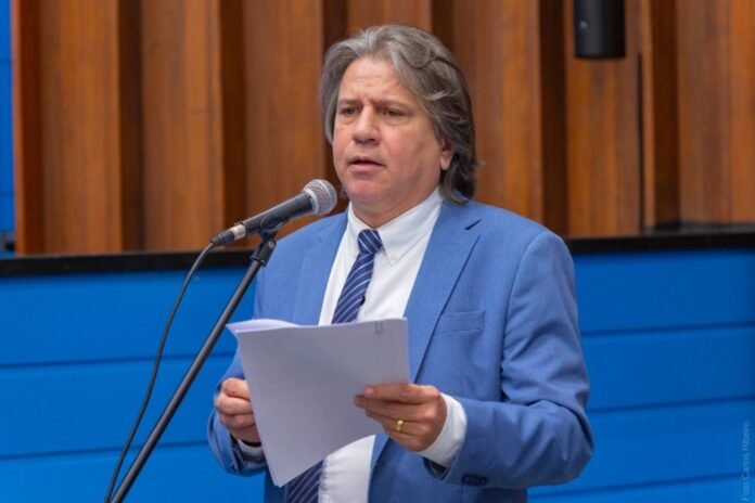 Man in a blue suit speaks into a microphone while holding papers in hand during a talk or presentation.
