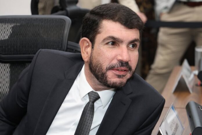Man in a dark suit and patterned tie sits at a conference table, looking to his right during a meeting.