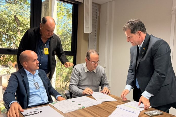 Four men in an office sign and review documents around a wooden table; one man in a suit stands while the others look on.