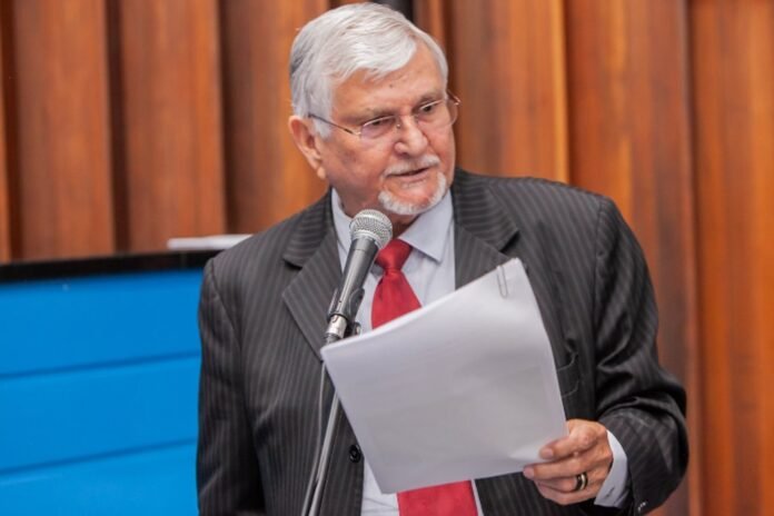 Older man in a pinstripe suit and red tie speaks into a microphone while holding papers at a podium-like setup.