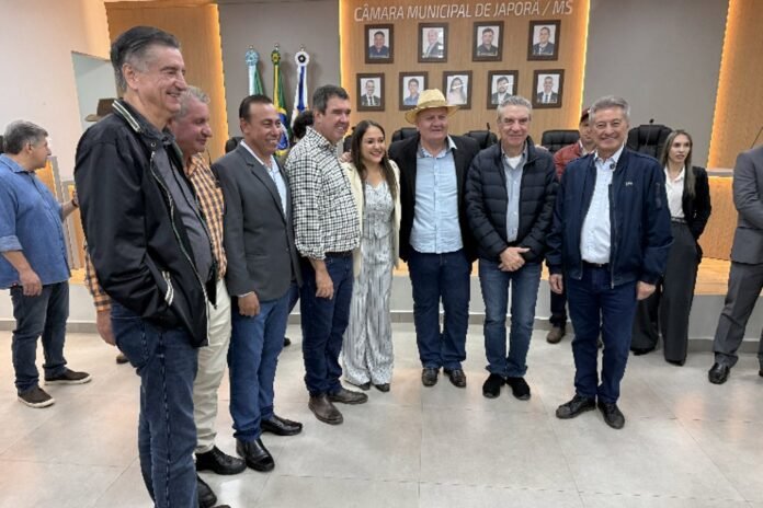 Group of officials and colleagues posing for a photo inside the Câmara Municipal de Japorã, MS, in front of a portrait wall and flags.