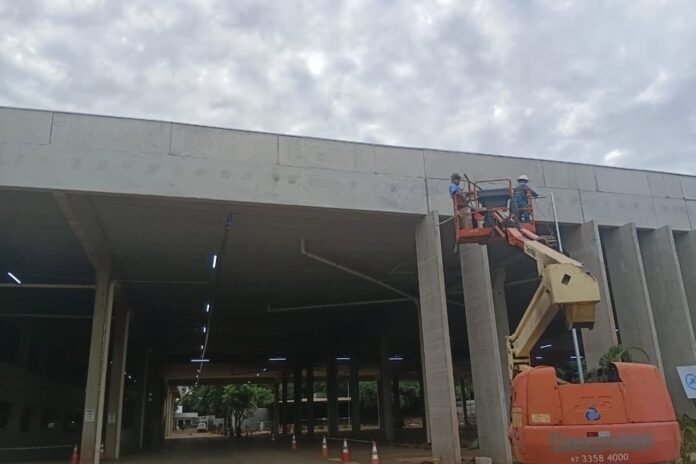 Two construction workers in hard hats stand in an elevated work platform beside a large gray concrete building under construction.