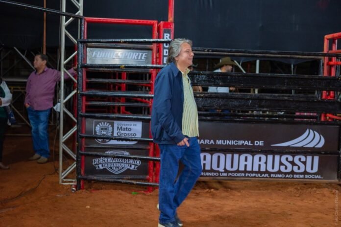 Man in a blue jacket and striped shirt walks beside a boxing ring at an outdoor event on a dirt arena surface.