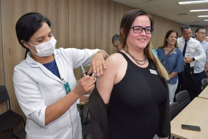 Healthcare worker gives a vaccination to a smiling woman in a black sleeveless top in a clinic, with others waiting in line behind them.