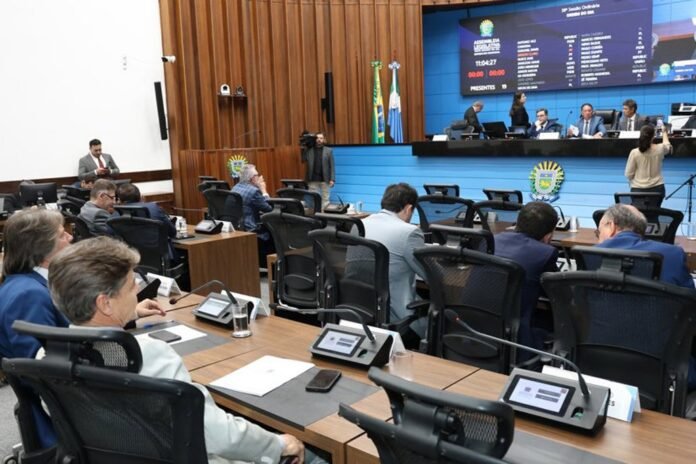 Parliament or council chamber with officials seated at desks, a raised blue dais, and a large digital screen showing data and scores in the background