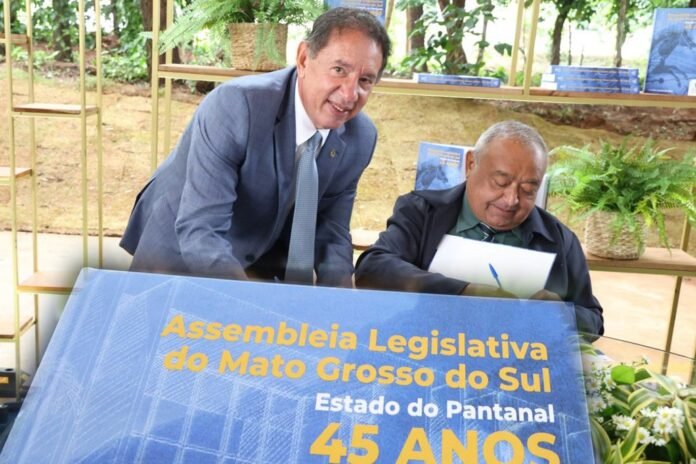 Two men in suits signing documents at a 45th anniversary event for the Mato Grosso do Sul Legislative Assembly in Brazil, with a blue banner in the foreground.