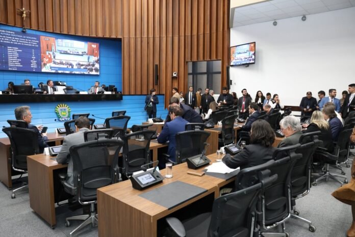 Parliamentary chamber with lawmakers seated at curved wooden desks, a blue panel backdrop, and a large screen displaying information at the front.