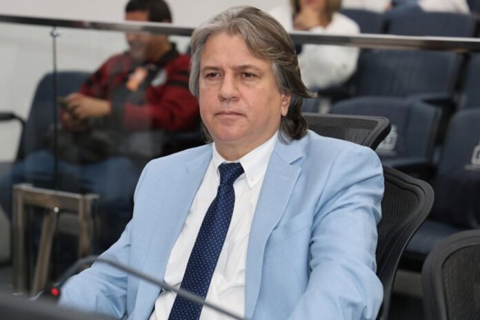 Man in a light blue suit and navy polka-dot tie seated at a conference table in a meeting room with a glass partition behind him.