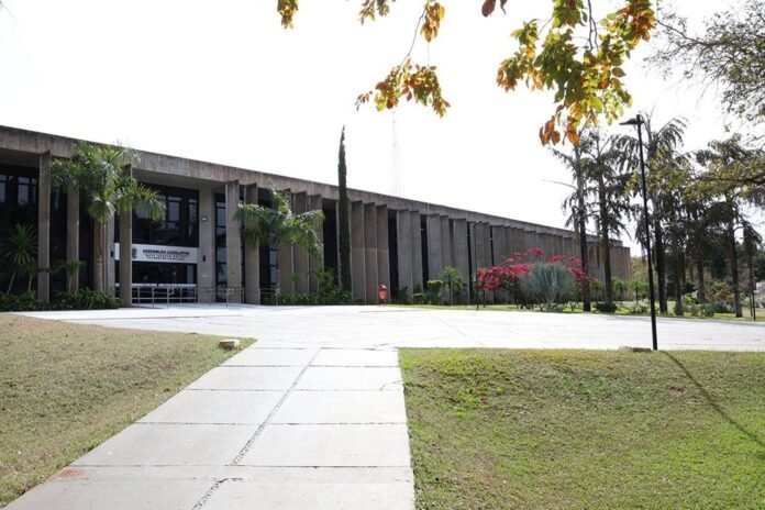 Exterior view of a modern government building with a long colonnade and concrete pillars, flanked by palm trees and a wide walkway toward the entrance (Assembleia Legislativa sign visible).
