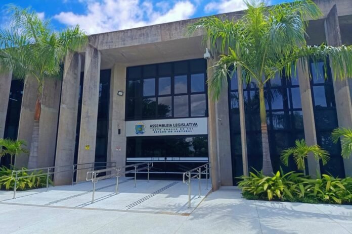 Front entrance of the Assembleia Legislativa building, with a ramp, glass doors, and palm trees.