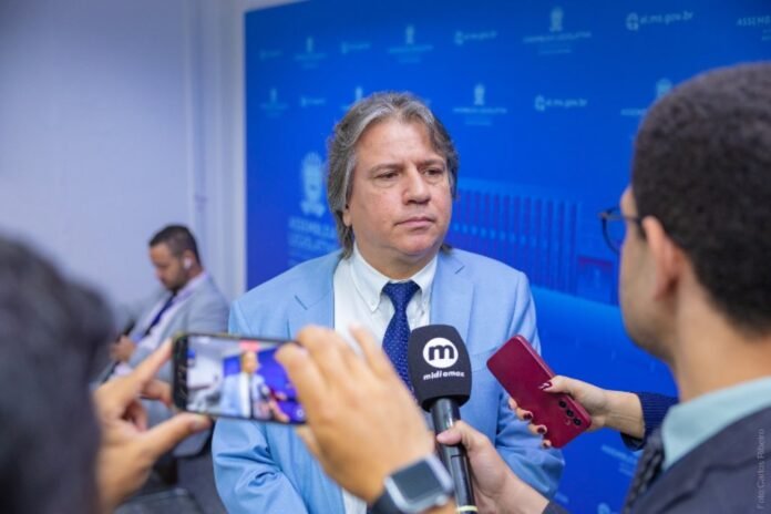 Man in a light blue suit giving an interview as reporters hold microphones and record with phones nearby.