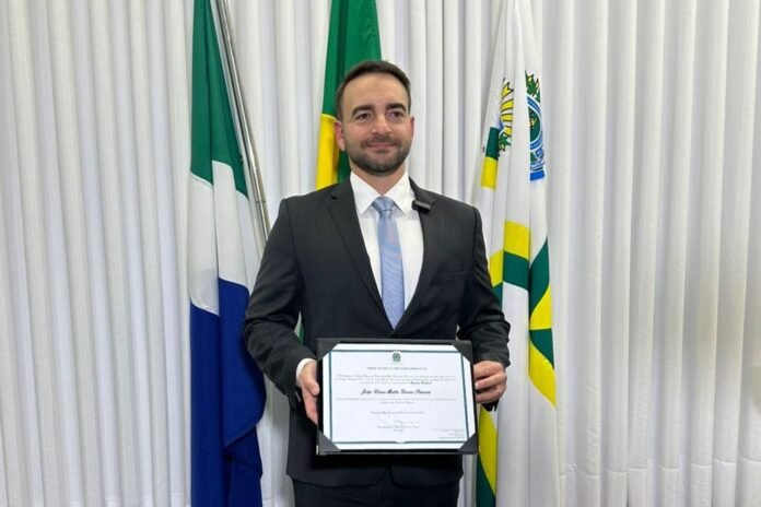 Man in a dark suit and light blue tie holds a framed certificate at a formal ceremony, with flags in the background.