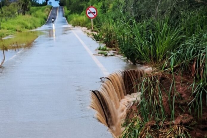 Rural road flooded with water spilling over a broken edge on the right, surrounded by tall grasses and a no-entry sign in the distance.
