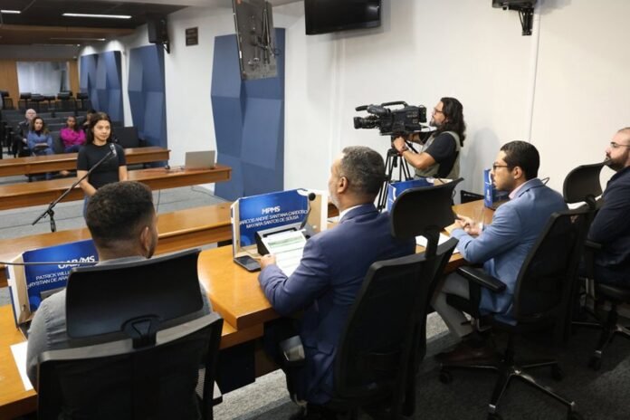 Young woman stands at a microphone speaking to a panel in a formal hearing room as a cameraman films the proceedings behind her.