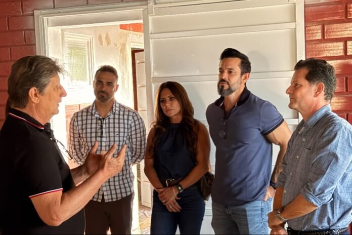 Five adults stand in a doorway listening as a man in a black polo speaks and gestures. The setting features a red brick wall and an open interior door behind them.