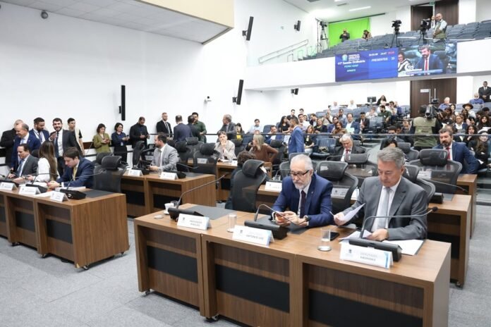 Officials seated at the front desks of a large conference room, nameplates and microphones in view, with a crowd behind them.