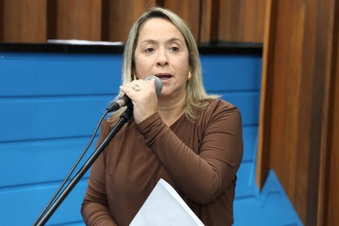 Woman in a brown long-sleeve top speaks into a handheld microphone while holding papers, in front of a blue wall backdrop.