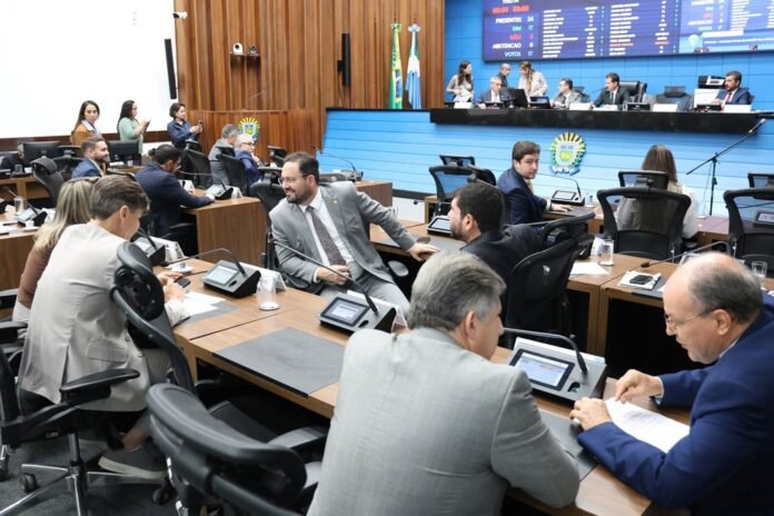 Council chamber scene with officials seated at desks with tablets and microphones, and a panel at the blue raised dais in the background.