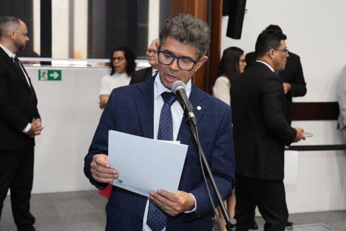 Man in a blue suit and glasses reads from a document at a microphone during a formal event.