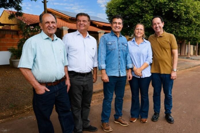 Five adults posing together outdoors on a sunny street, smiling at the camera with casual attire.
