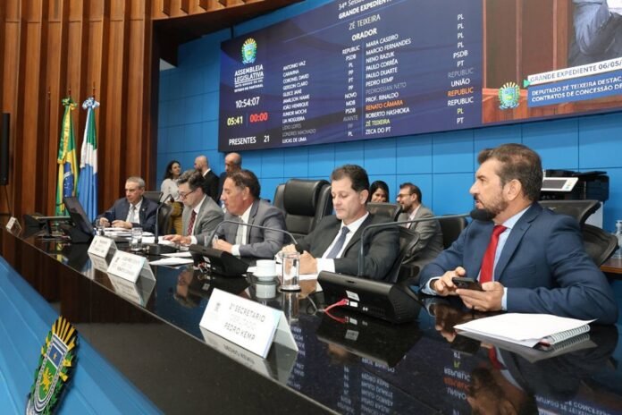 Parliamentary panel in a blue chamber; suited officials sit at a curved desk with microphones and nameplates.