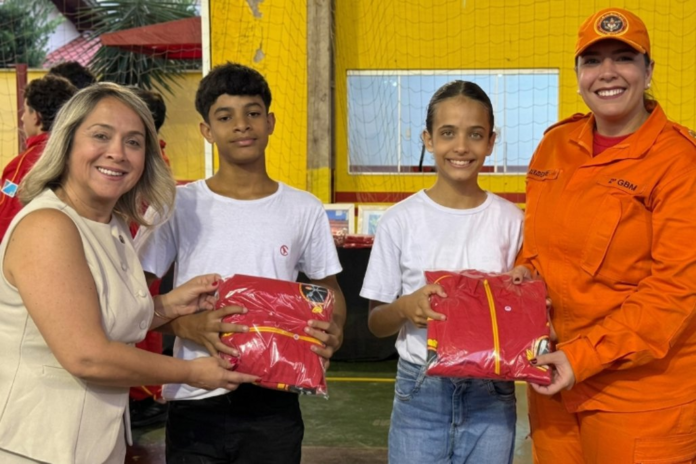 Woman in a white sleeveless top and an orange-uniformed person hand red packaged gifts to two kids in white shirts during a donation event. The scene is indoors with a yellow wall in the background.