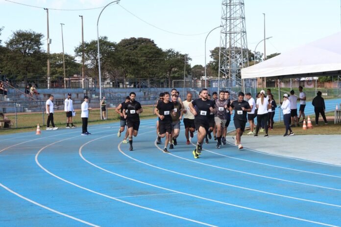 A group of runners at the start of a race on a bright blue outdoor track with onlookers nearby under a white tent.