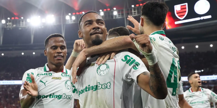 Palmeiras players celebrate a goal, embracing in white jerseys with green logos in a packed stadium.