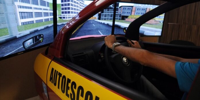 Person sits in a red and yellow car cockpit, hands on a steering wheel, driving in a multi-screen driving simulator setup with an urban road scene.
