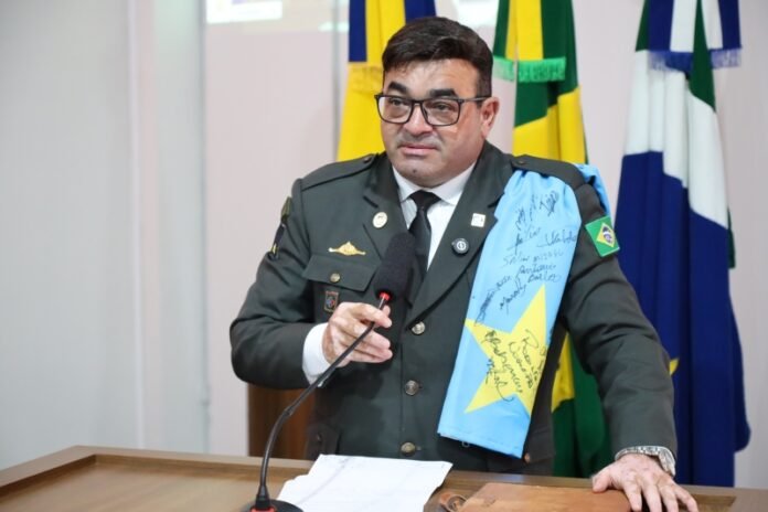 Military officer speaks at a podium with a microphone; a signed blue sash rests over his shoulder, with flags in the background.