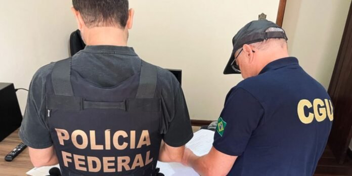 Two Brazilian law enforcement officers, one in a Polícia Federal vest and the other in a CGU shirt, review papers at a desk.