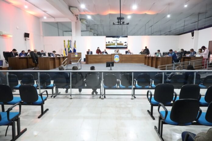 A formal council or board meeting in a conference hall with officials seated at long desks facing the audience, flags in the background, and a large screen at the back.