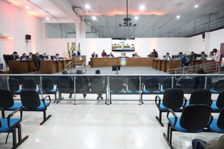 A formal council or board meeting in a conference hall with officials seated at long desks facing the audience, flags in the background, and a large screen at the back.