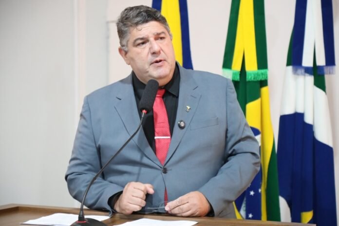 Man in a gray suit and red tie speaking into a microphone at a podium, with colorful flags in the background