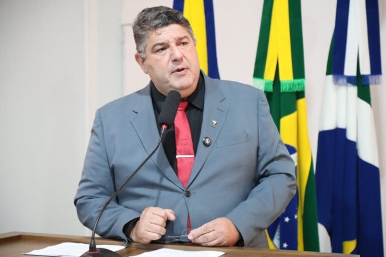 Man in a gray suit and red tie speaking into a microphone at a podium, with colorful flags in the background