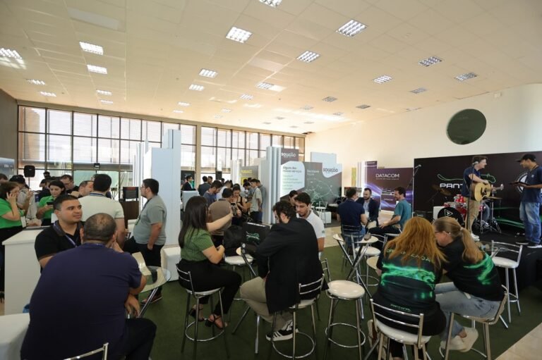 Crowd at an indoor tech expo with booths, people chatting at tables, and a live band performing on stage in the background.