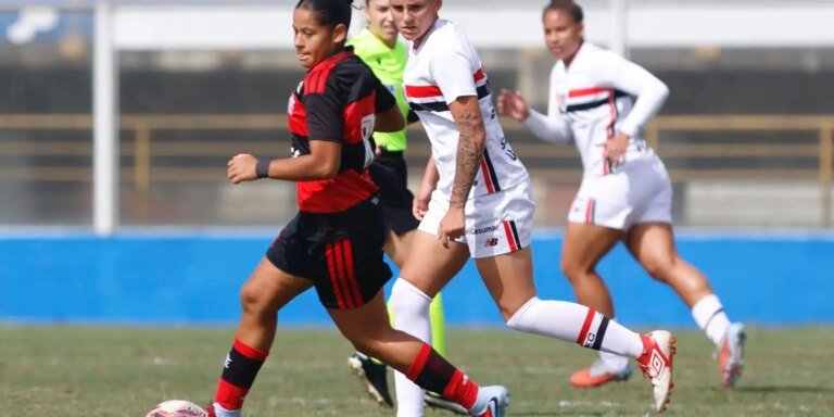 Soccer players sprint for the ball on a green field during a match; one in red-and-black kit leading ahead of white kits with a tattooed arm visible on a white-uniformed player.