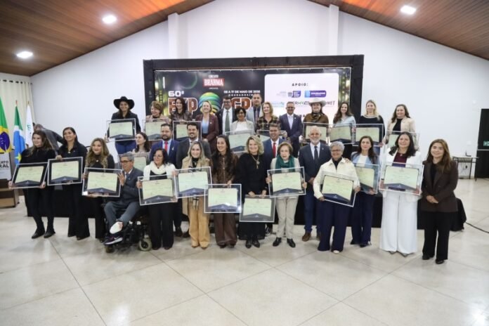 Group of men and women posing with certificates at an awards ceremony in a hall; a large, diverse group stands in front of a stage banner with flags nearby.