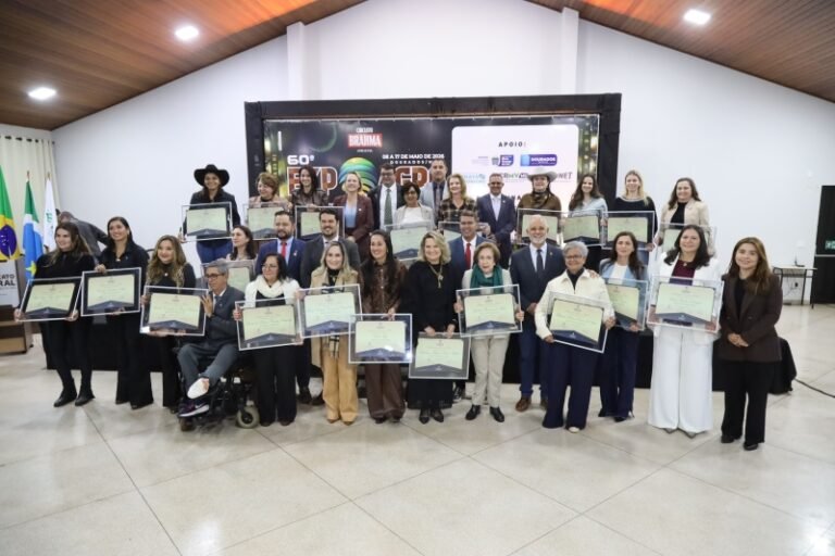 Group of men and women posing with certificates at an awards ceremony in a hall; a large, diverse group stands in front of a stage banner with flags nearby.
