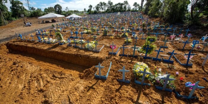Wide view of a graveyard with dozens of blue crosses marking graves, dirt ground, and flowers; white tents and trees in the background.