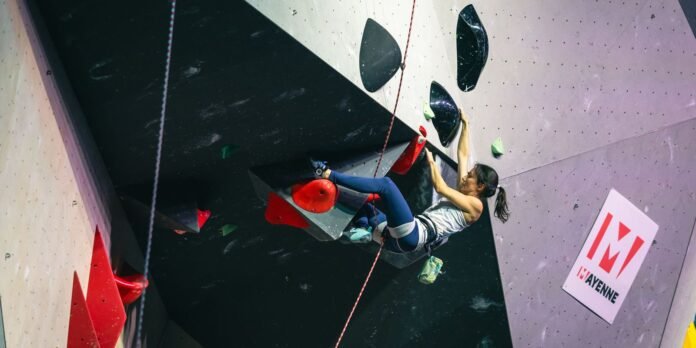 Female climber wearing a harness and chalk bag climbs an indoor wall, gripping a red hold with her left and a black hold with her right on an overhanging route.