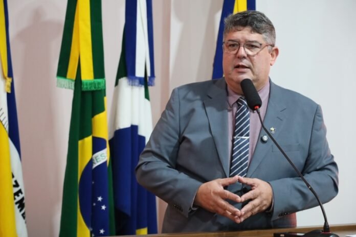 Man in a gray suit and striped tie giving a speech at a podium with a microphone, flags in the background.